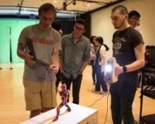 Students examining a small humanoid robot displayed on a pedestal in a laboratory or studio setting.