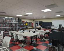 Makerspace classroom with collaborative work tables, 3D printers, prototyping equipment, and a large display screen.