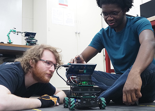 Copilot said: Two people testing and adjusting a small mobile robot with onboard electronics in a robotics laboratory.