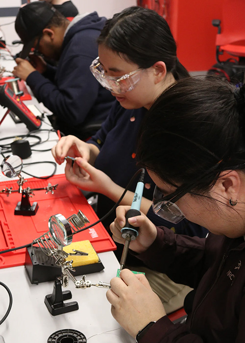People using soldering tools and electronic components at a workstation during a hands-on electronics and engineering lab activity.